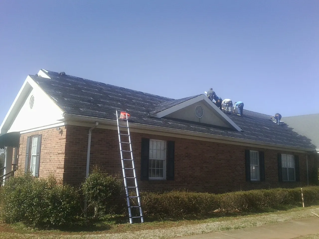 Crew tearing shingles off a brick ranch roof with a ladder set — Emergency Roof Repair work by our crew in Lawler
