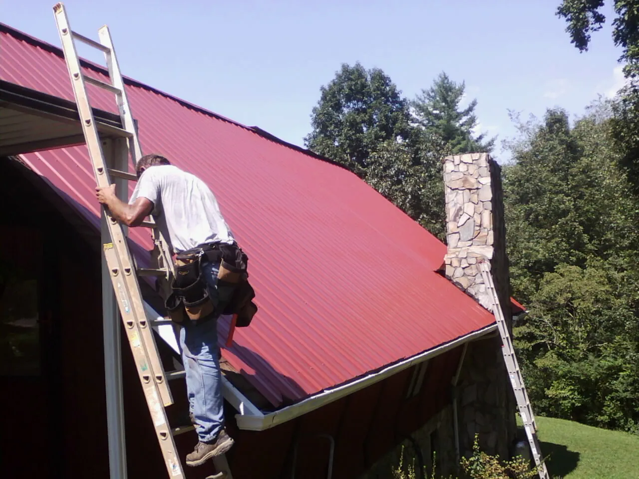 Roofer on a ladder beside a red metal roof and stone chimney — Soffit & Fascia Repair by Roofing Brothers in Lawler